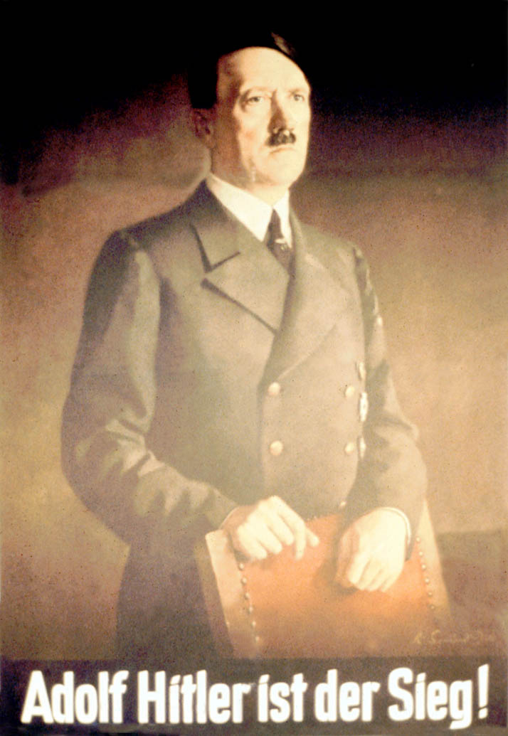 Hitler stands in a double breasted suit behind the back of a chair